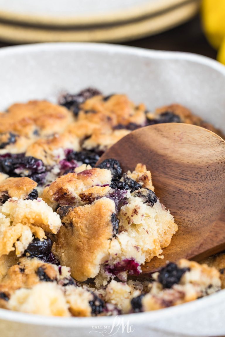 A wooden spoon scoops out a portion of blueberry cobbler from a white dish, showing golden-brown biscuit topping and cooked blueberries.