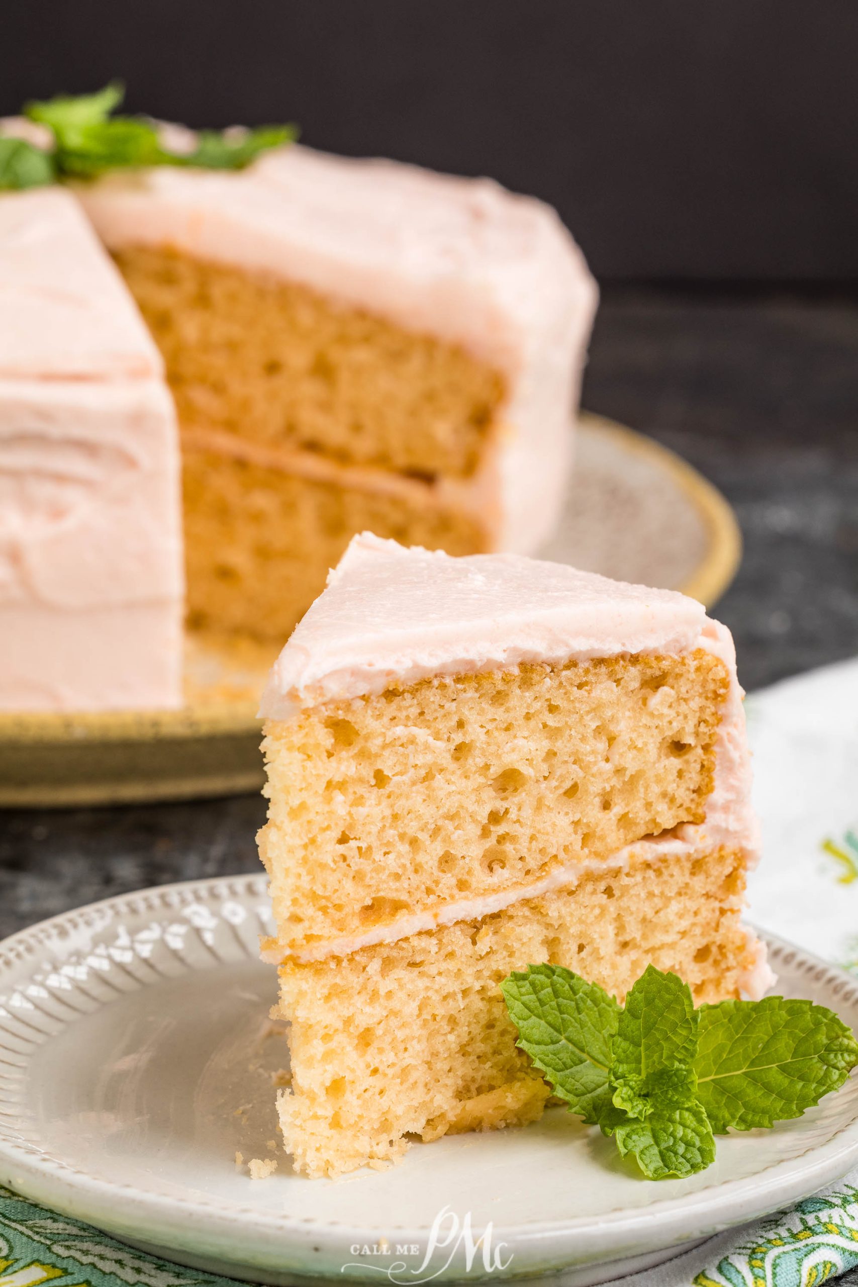 A slice of Pink Champagne Layer Cake with pink frosting sits on a plate, garnished with a sprig of mint; the rest of the cake is in the background.