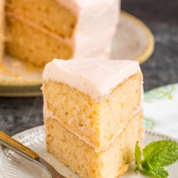 A slice of Pink Champagne Layer Cake with light pink frosting sits on a white plate, garnished with a mint sprig and a fork.