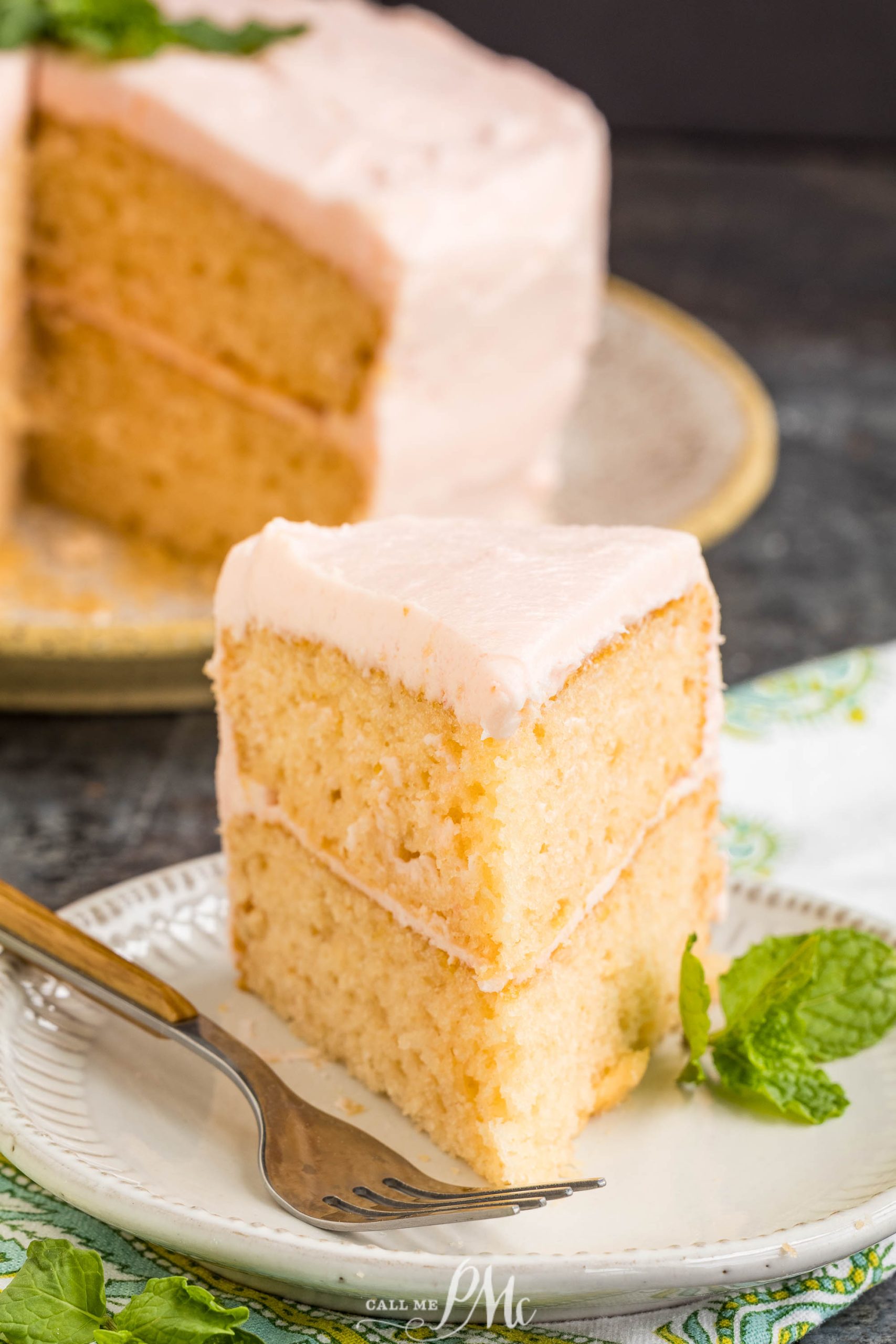 A slice of dessert with light pink frosting sits on a white plate, garnished with a mint sprig and a fork.