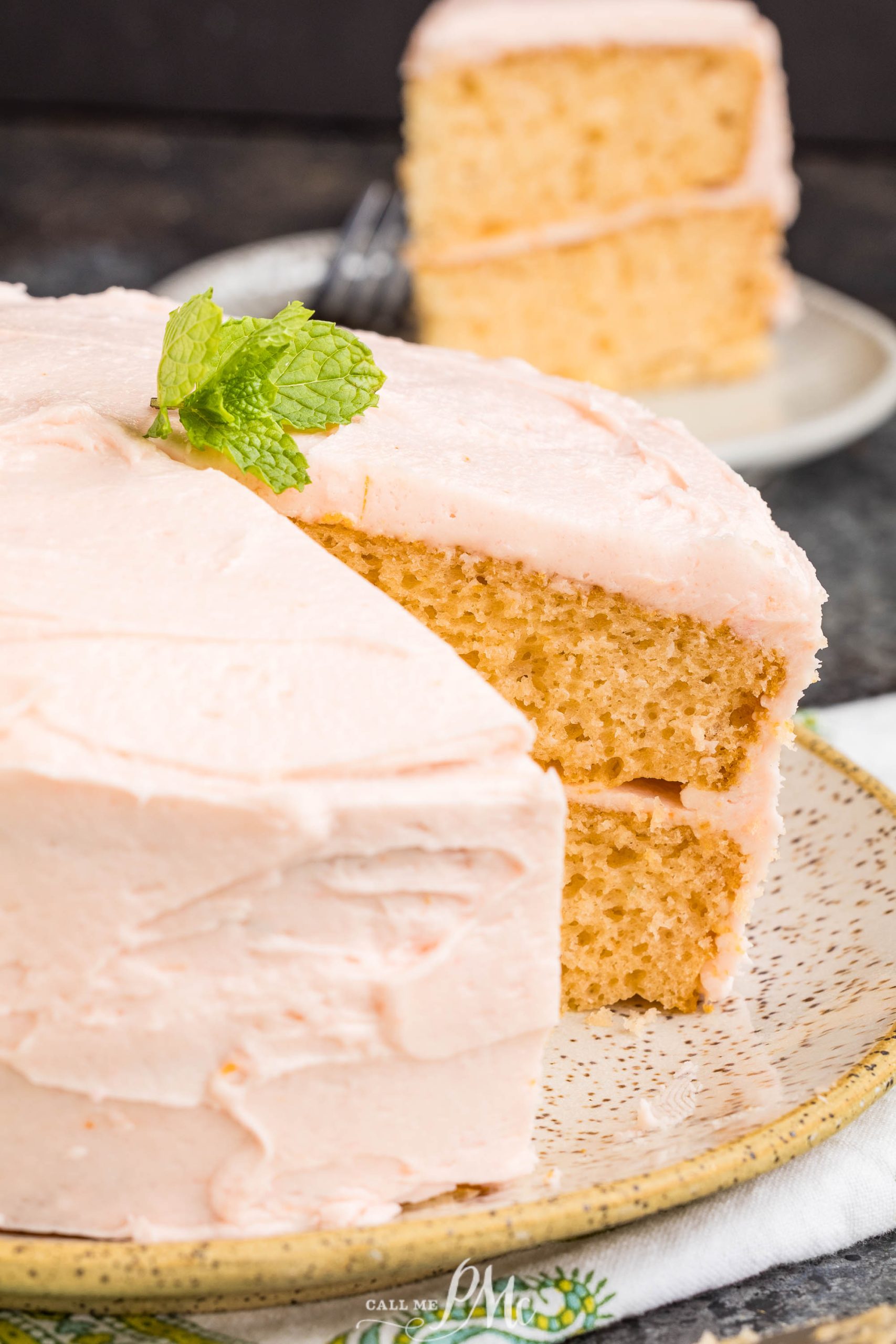 A dessert featuring two vanilla layers with light pink frosting and a mint garnish, with one slice cut and served on a plate in the background.
