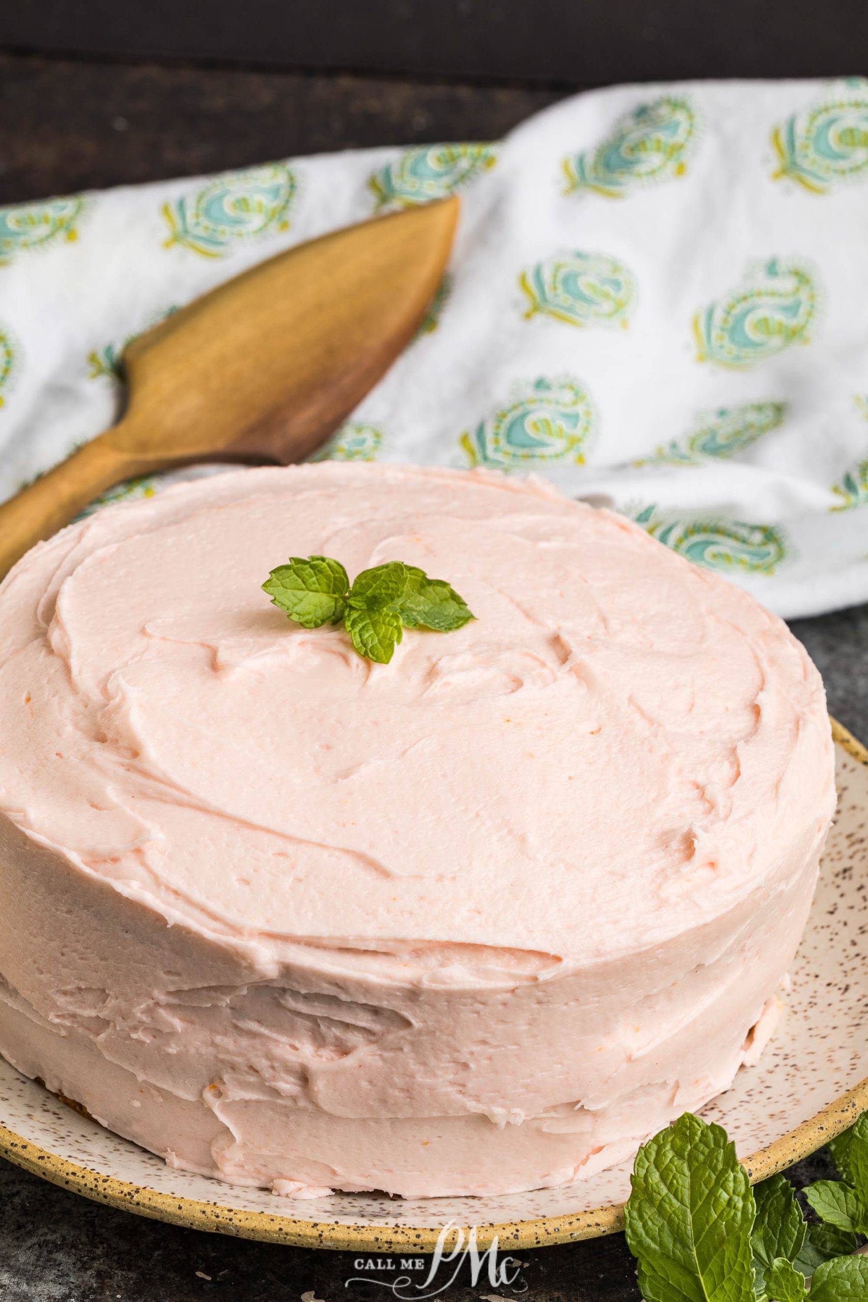 A Pink Champagne Layer Cake with smooth pink frosting, topped with a sprig of mint, sits on a speckled plate next to a wooden spatula and patterned cloth.