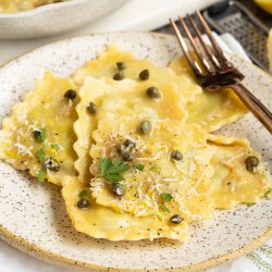 A plate of ravioli topped with grated cheese, capers, herbs, and a fork on the side.