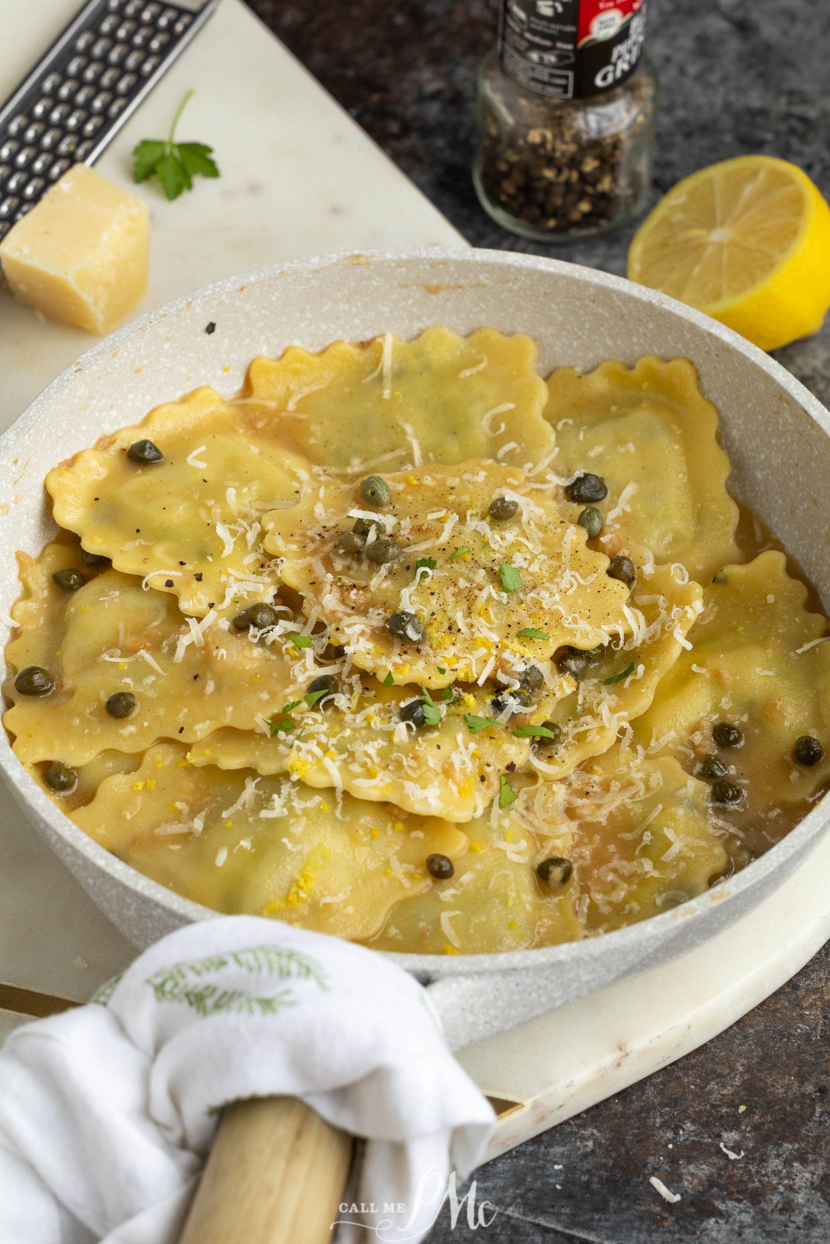 A skillet of pasta topped with grated cheese, capers, and herbs sits on a countertop with a lemon half, pepper grinder, and cheese grater nearby.