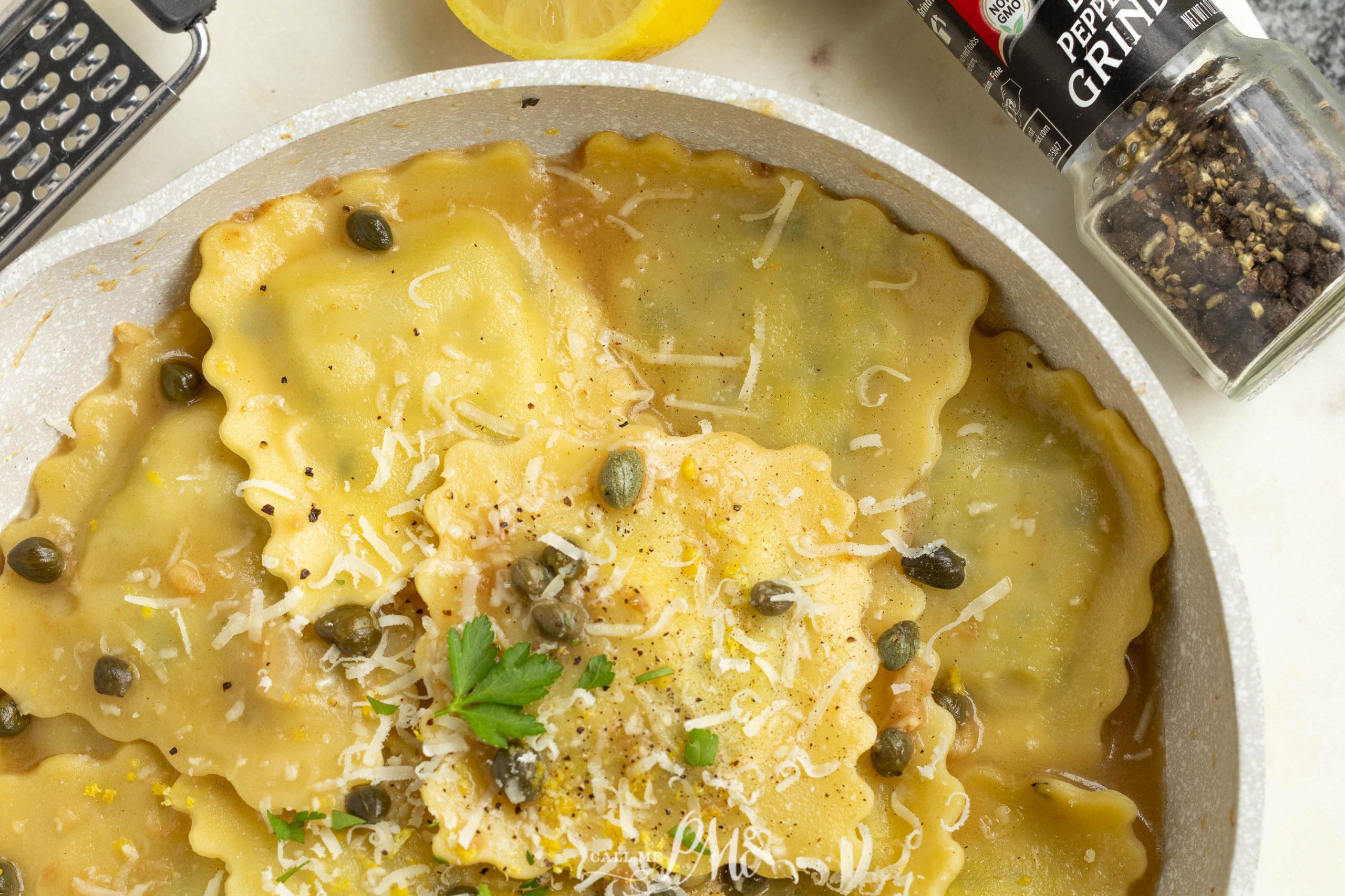 A plate of ravioli topped with grated cheese, capers, parsley, and lemon zest sits next to a pepper grinder and a lemon half.