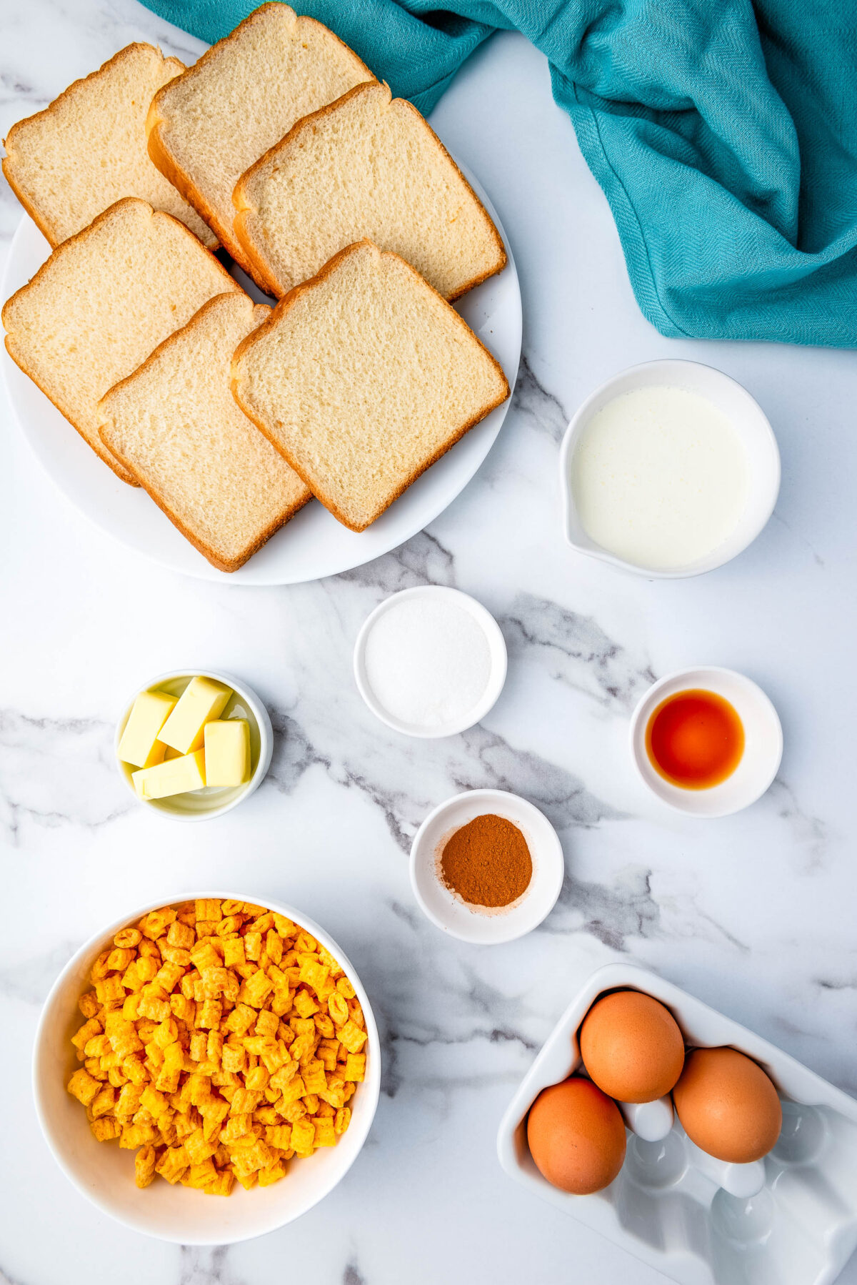 Slices of bread on a plate, a bowl of yellow cereal, cubes of butter, eggs in a carton, small bowls of sugar, cinnamon, vanilla extract, and a jug of milk, all arranged on a white marble surface with a blue cloth.