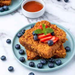 Two slices of Cap'n Crunch French Toast topped with sliced strawberries, blueberries, and a mint sprig on a blue plate. Extra blueberries are scattered around, with a cup of syrup and strawberries in the background on a marble surface.