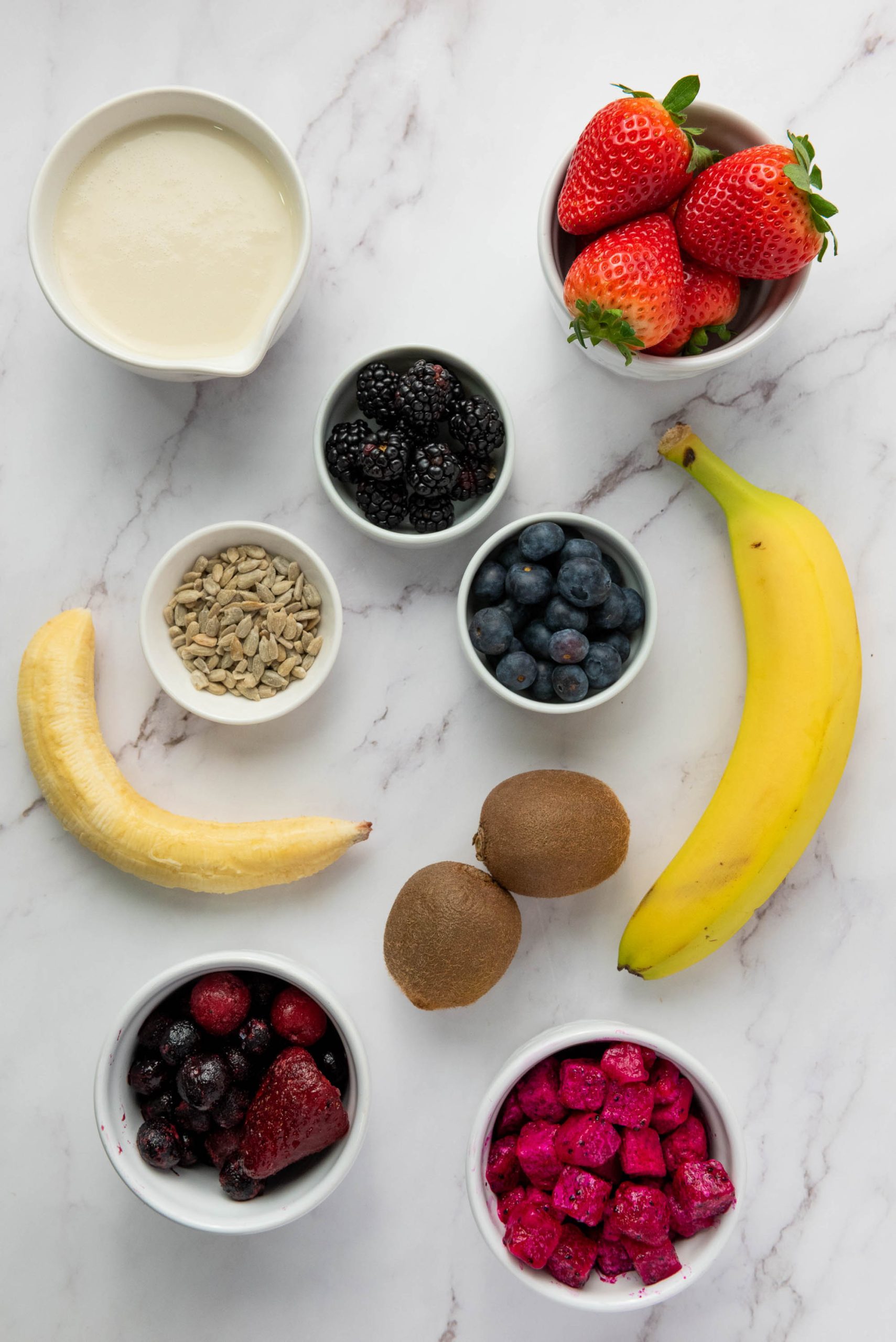Assorted fruits, berries, sunflower seeds, and a cup of yogurt arranged on a white marble surface.
