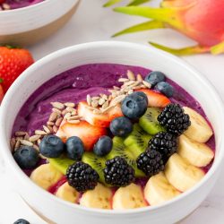 A dragon fruit smoothie bowl topped with sliced bananas, kiwi, strawberries, blackberries, blueberries, and sunflower seeds, with extra dragon fruit and strawberries in the background.