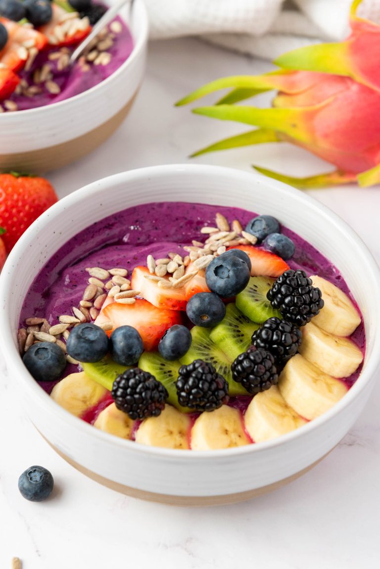 A dragon fruit smoothie bowl topped with sliced bananas, kiwi, strawberries, blackberries, blueberries, and sunflower seeds, with extra dragon fruit and strawberries in the background.