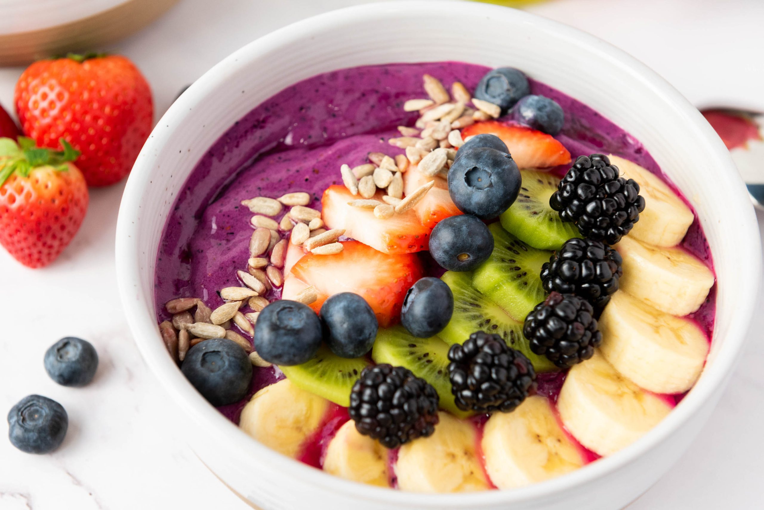 A bowl of pink topped with sliced banana, kiwi, strawberries, blackberries, blueberries, and sunflower seeds, with a few berries on the side.
