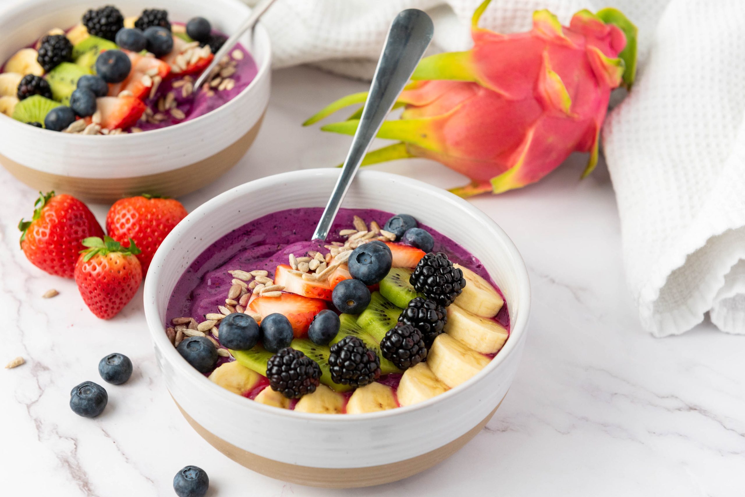 A vibrant dragon fruit smoothie bowl topped with sliced banana, kiwi, strawberries, blueberries, blackberries, and sunflower seeds, with a spoon and fresh dragon fruit in the background.