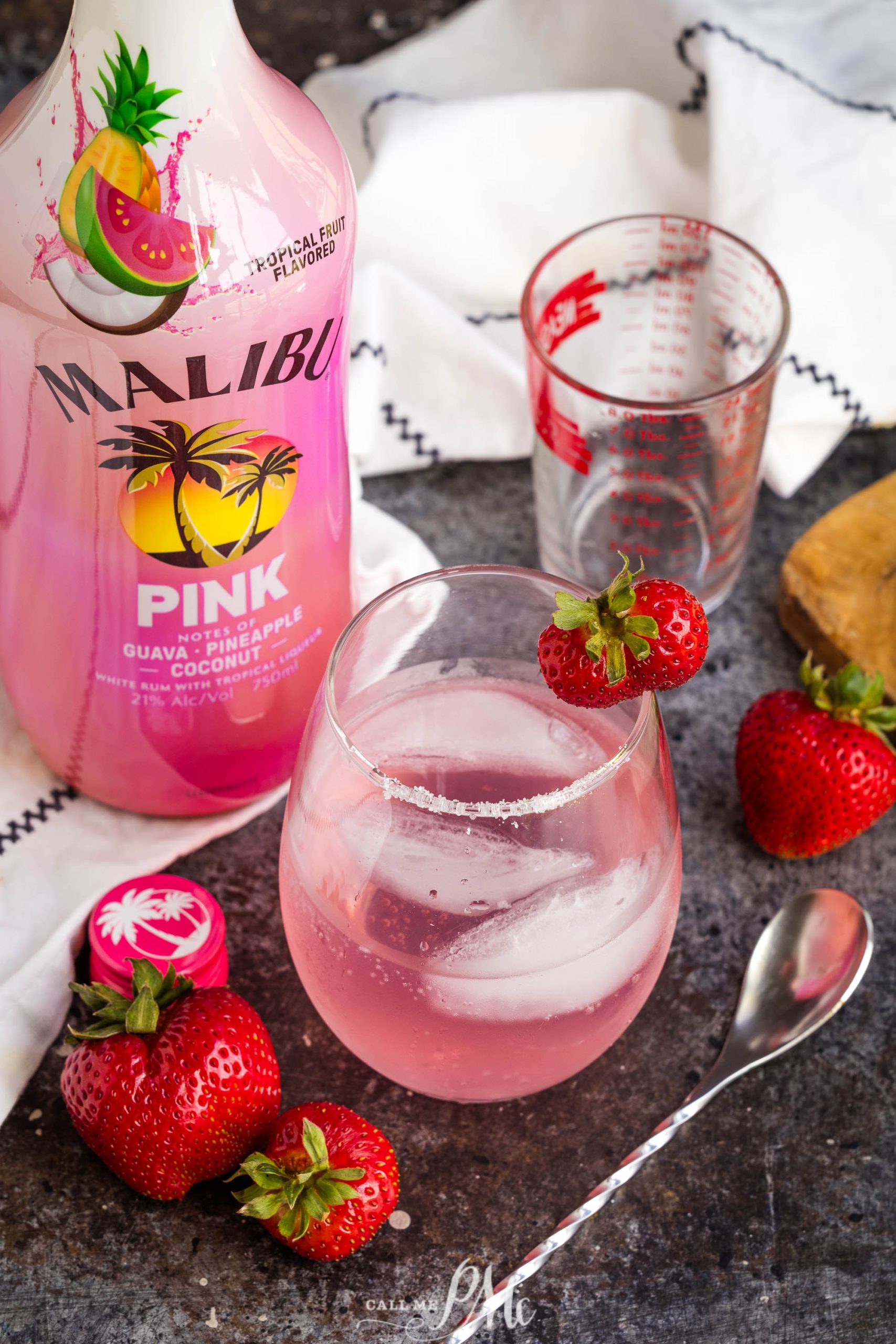 A cocktail with ice and a strawberry garnish sits next to a liquor bottle, fresh strawberries, a measuring cup, and a spoon on a dark countertop.