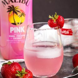 A glass of pink cocktail garnished with a strawberry, with fresh strawberries, a bottle of Malibu Pink, and a measuring cup in the background.