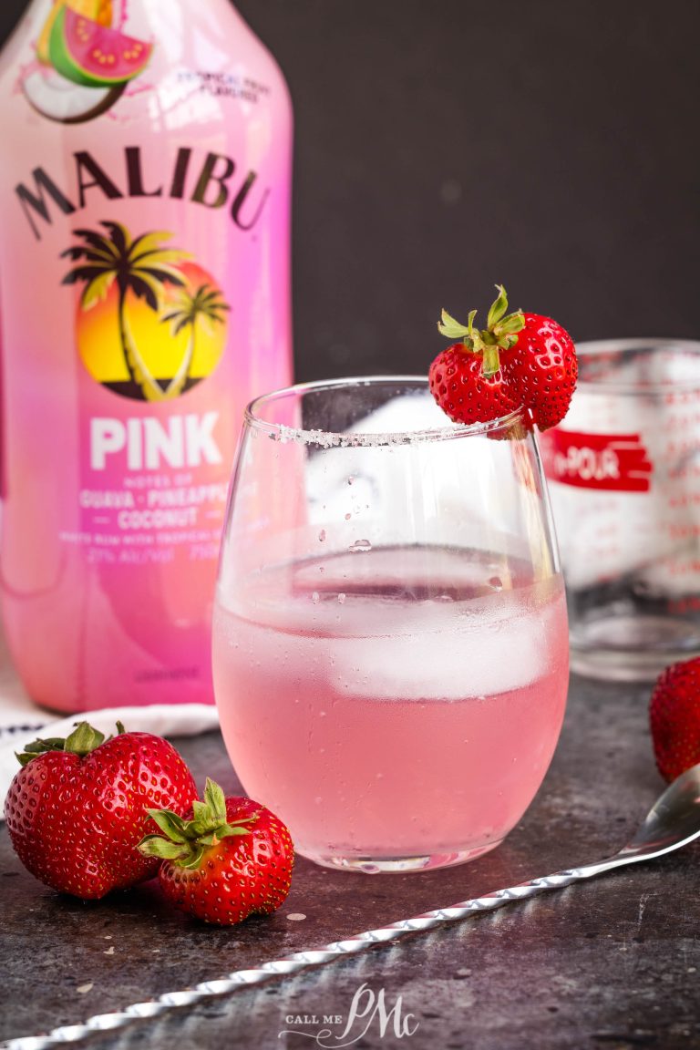 A glass of pink cocktail garnished with a strawberry, with fresh strawberries, a bottle of Malibu Pink, and a measuring cup in the background.