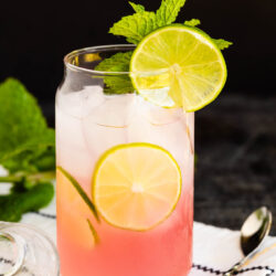 A glass of Malibu Pink Limeade with ice, garnished with lime slices and fresh mint, sits on a white cloth napkin with a silver spoon nearby. The drink showcases a beautiful pink-to-clear gradient from bottom to top.