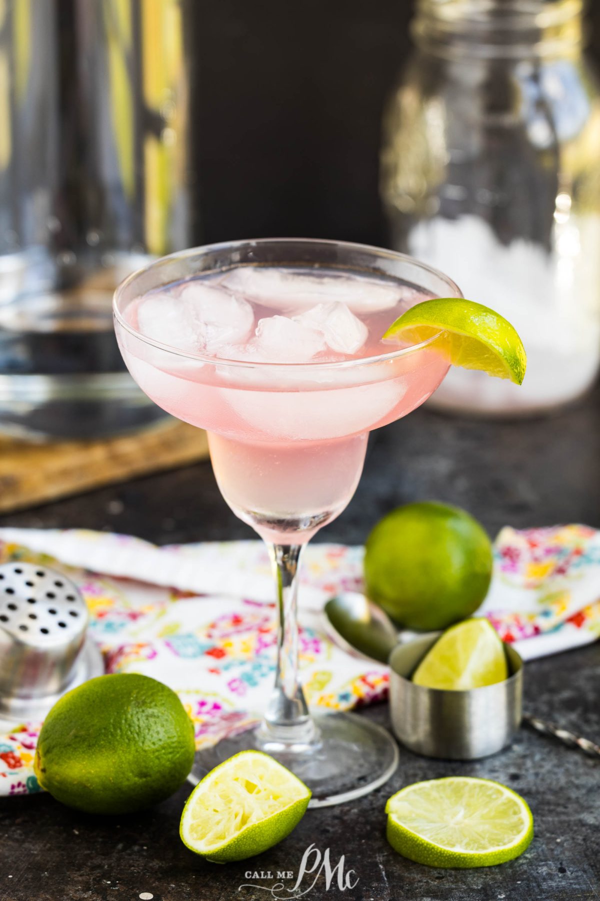 A pink cocktail with ice and a lime wedge is served in a margarita glass, surrounded by whole and sliced limes, a floral napkin, and a metal jigger on a dark surface.