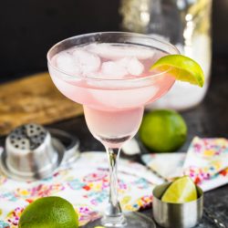 A pink cocktail with ice and a lime wedge in a margarita glass, surrounded by limes, a cocktail shaker, and a mason jar on a colorful napkin.