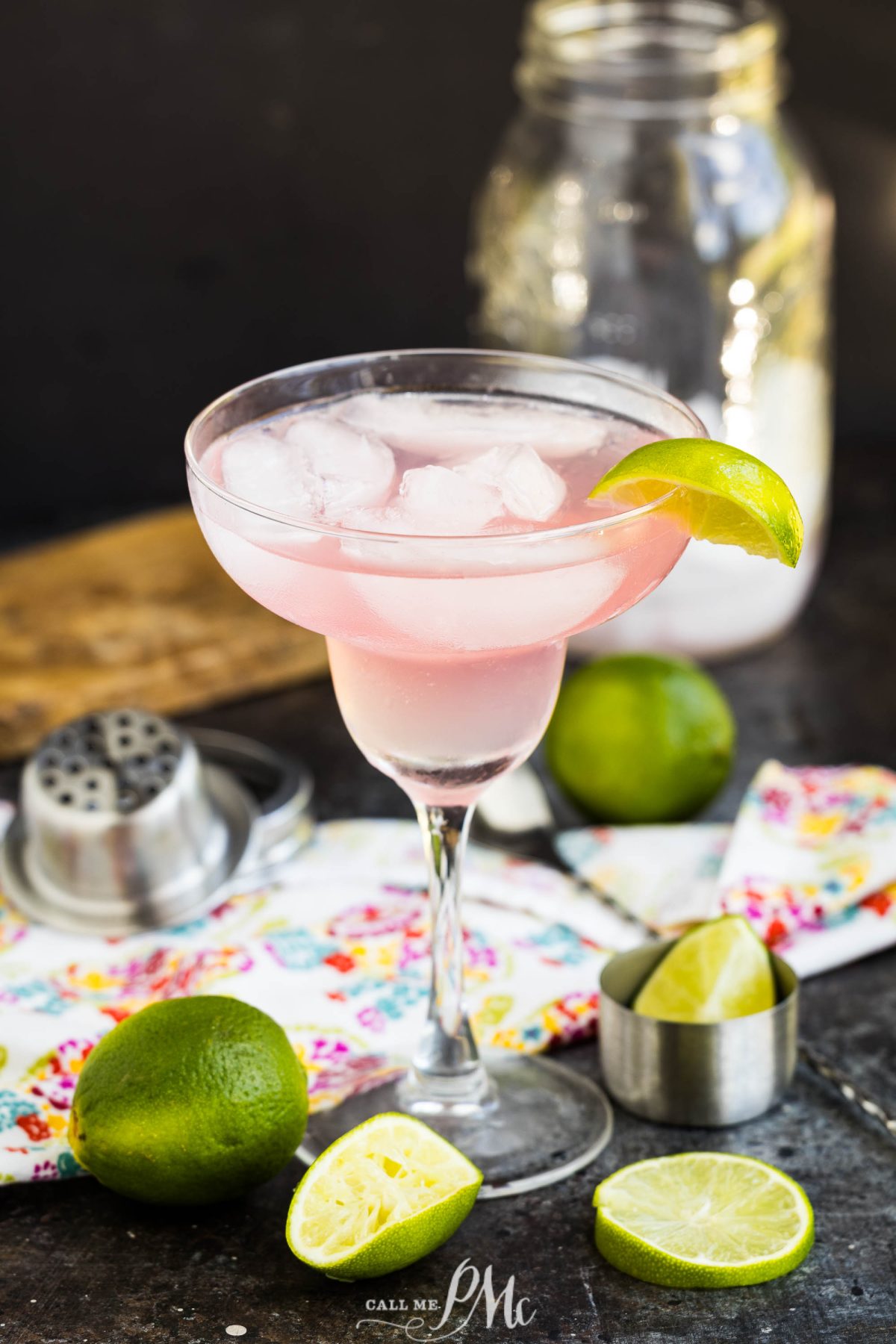 A cocktail with ice and a lime wedge in a margarita glass, surrounded by limes, a cocktail shaker, and a mason jar on a colorful napkin.