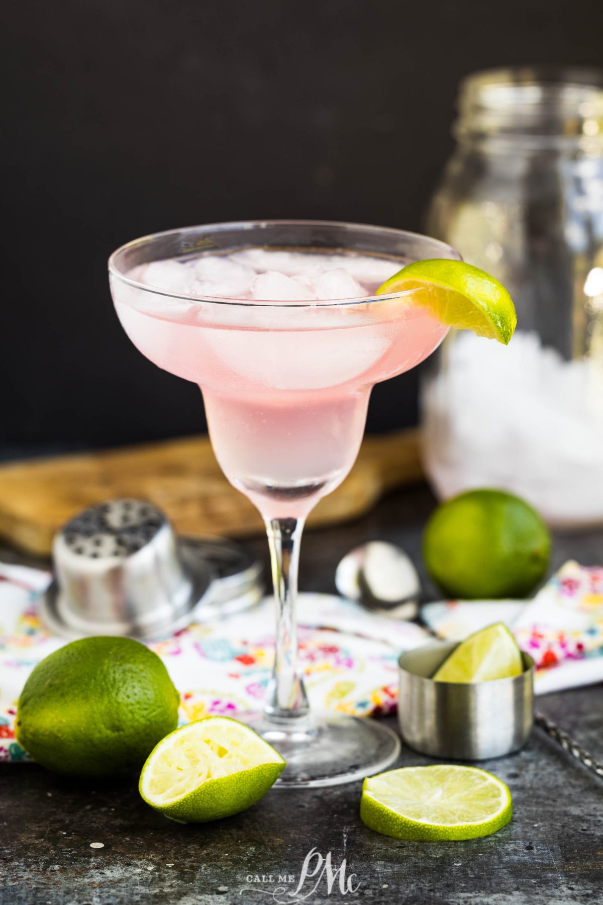 A cocktail with ice is served in a cocktail glass, garnished with a lime wedge. Fresh limes, a jigger, and a shaker are nearby on a colorful cloth.