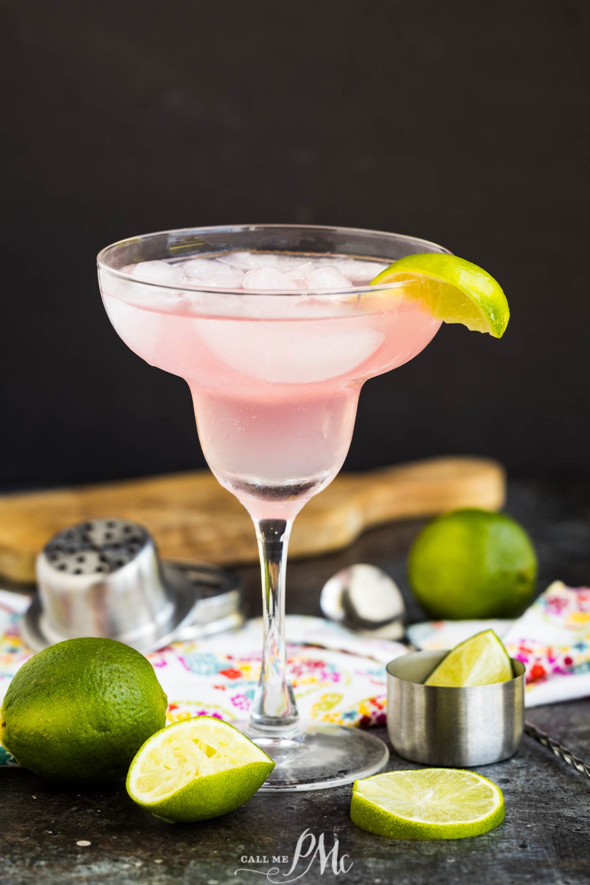 A pink margarita in a glass with ice and a lime wedge garnish, surrounded by fresh limes, a jigger, and a citrus squeezer on a dark surface.