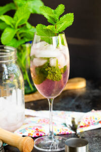 A wine glass filled with Malibu Pink Limeade, ice cubes, fresh mint leaves, and topped with a mint sprig. A mason jar, muddler, colorful napkin, and mint plant are in the background.