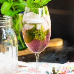 A wine glass filled with Malibu Pink Limeade, ice cubes, fresh mint leaves, and topped with a mint sprig. A mason jar, muddler, colorful napkin, and mint plant are in the background.
