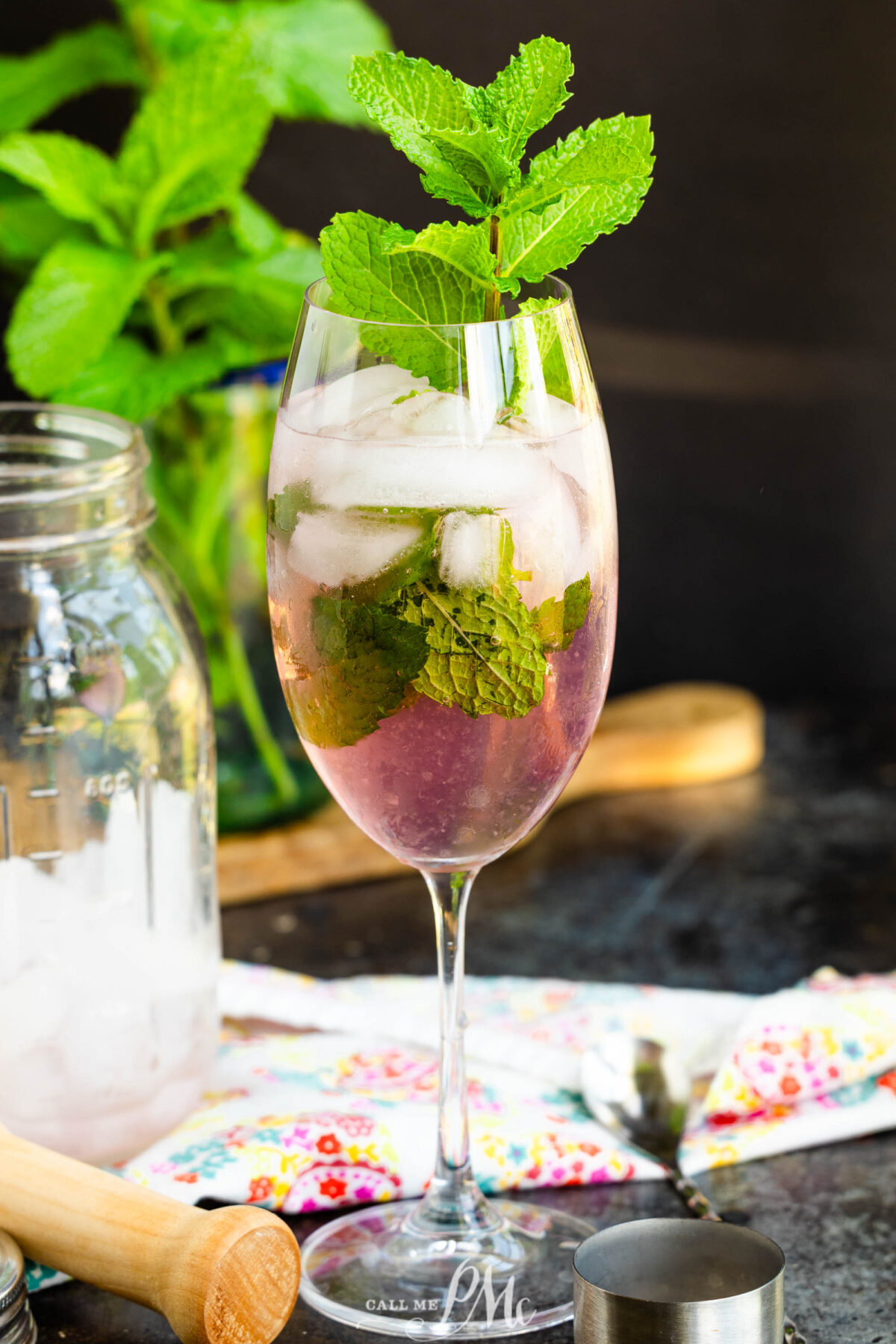 A wine glass drink, ice cubes, fresh mint leaves, and topped with a mint sprig. A mason jar, muddler, colorful napkin, and mint plant are in the background.