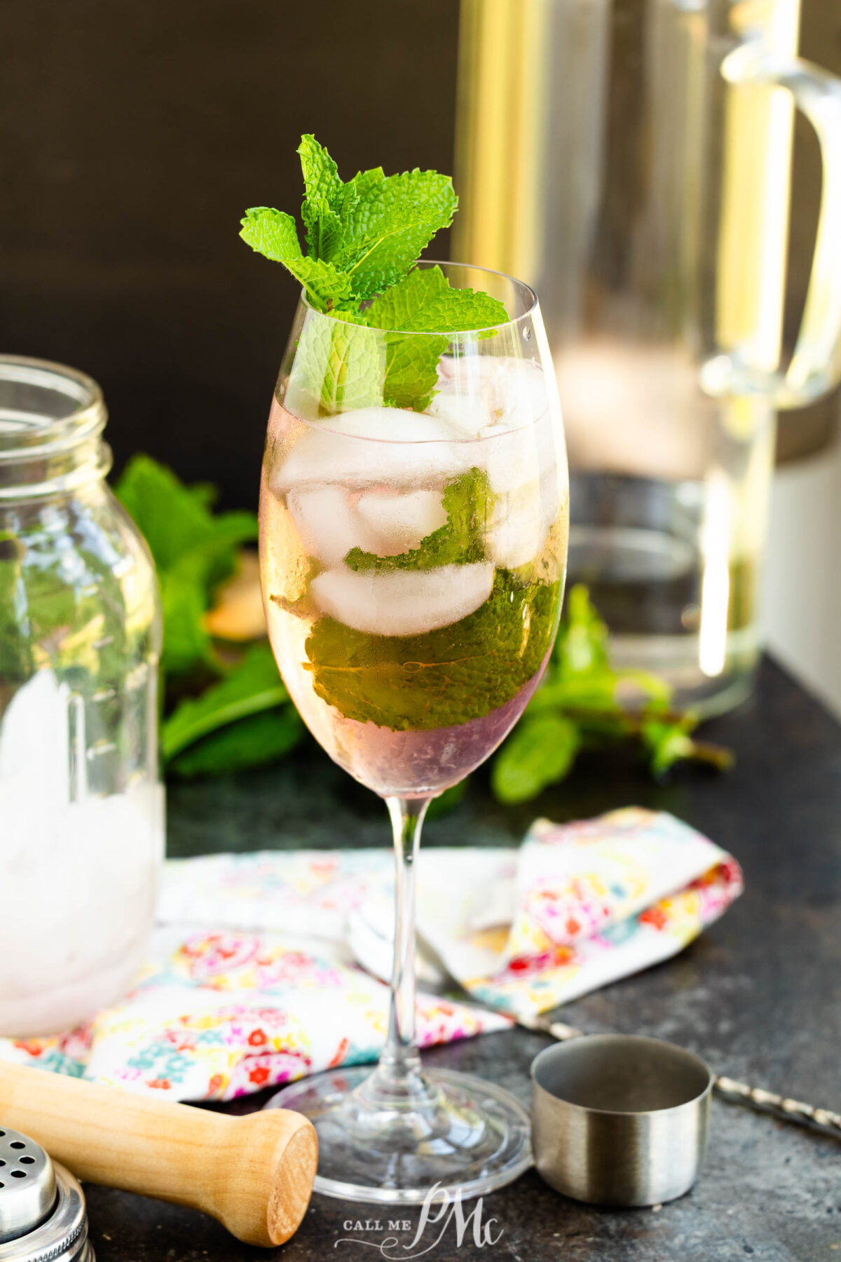 A tall wine glass filled with a minty Malibu Pink Limeade, garnished with fresh mint leaves. The glass sits on a dark countertop alongside a mason jar, colorful napkin, jigger, and muddler.
