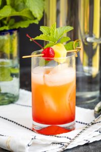 A tall glass filled with a vibrant Malibu Pink Pineapple Sunset Cocktail, ice, and garnished with a cherry, pineapple slice, and mint sprig on a napkin.