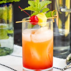 A tall glass filled with a vibrant Malibu Pink Pineapple Sunset Cocktail, ice, and garnished with a cherry, pineapple slice, and mint sprig on a napkin.