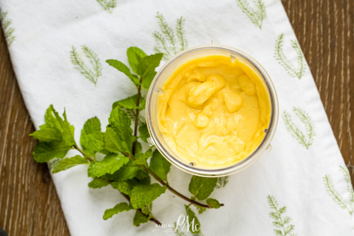 A blender cup filled with creamy yellow mango ice cream sits on a white cloth with green leaf patterns, next to a sprig of fresh mint on a wooden surface.