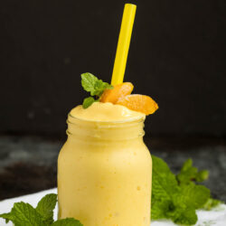 A mason jar filled with a creamy yellow smoothie, garnished with peach slices and mint leaves, sits on a white cloth. A yellow straw is placed in the drink, and fresh mint surrounds the jar. The background is dark.