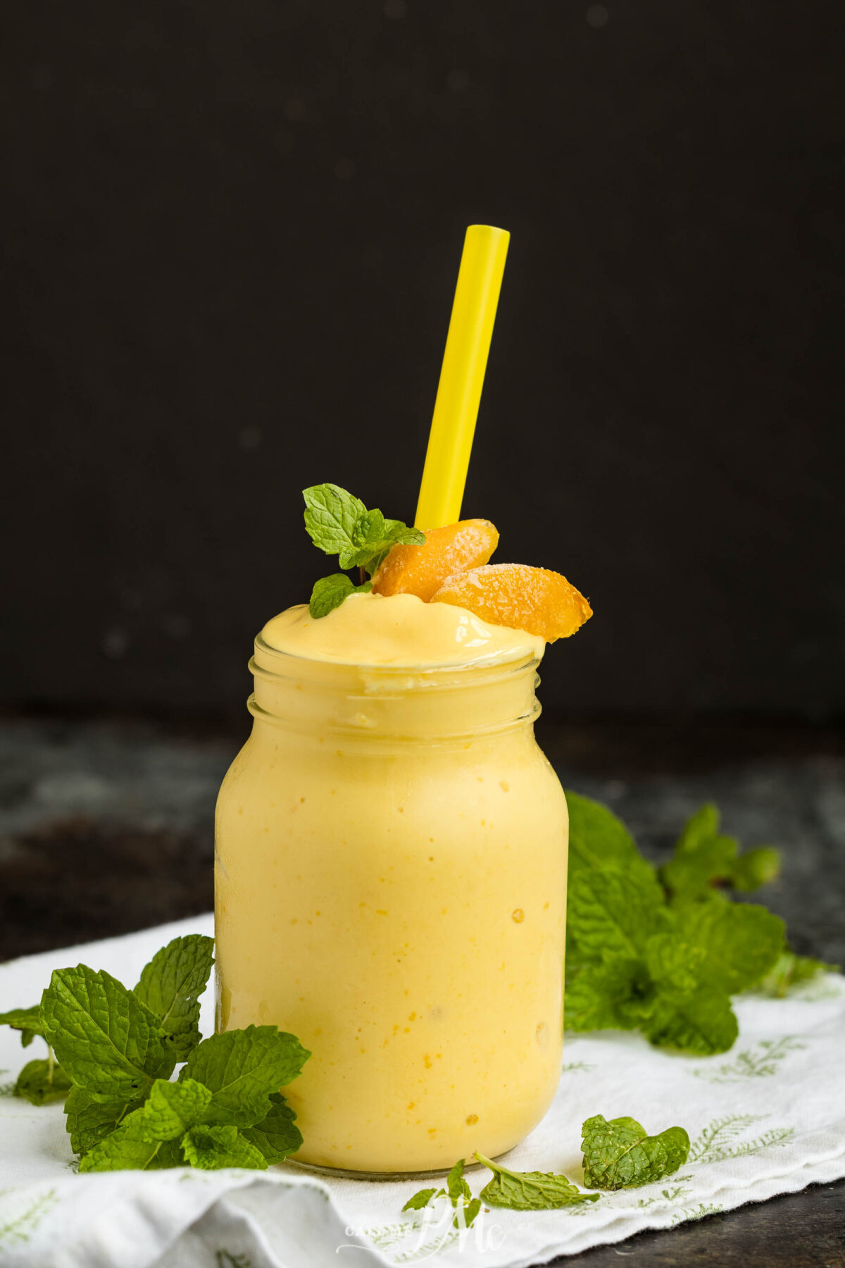 A mason jar filled with a creamy yellow smoothie, garnished with peach slices and mint leaves, sits on a white cloth. A yellow straw is placed in the drink, and fresh mint surrounds the jar. The background is dark.