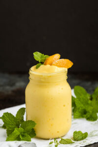 A mason jar filled with creamy yellow mango smoothie, garnished with mango slices and fresh mint leaves, sits on a white cloth with additional mint sprigs nearby. The background is dark and blurred.