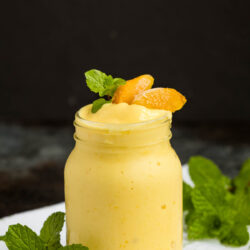 A mason jar filled with creamy yellow mango smoothie, garnished with mango slices and fresh mint leaves, sits on a white cloth with additional mint sprigs nearby. The background is dark and blurred.