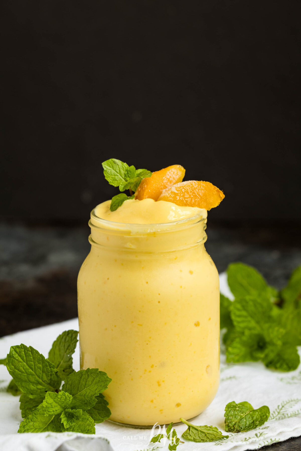 A mason jar filled with creamy yellow mango smoothie, garnished with mango slices and fresh mint leaves, sits on a white cloth with additional mint sprigs nearby. The background is dark and blurred.
