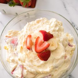 A glass bowl filled with creamy fruit salad topped with sliced strawberries and bananas, with a container of fresh strawberries in the background.