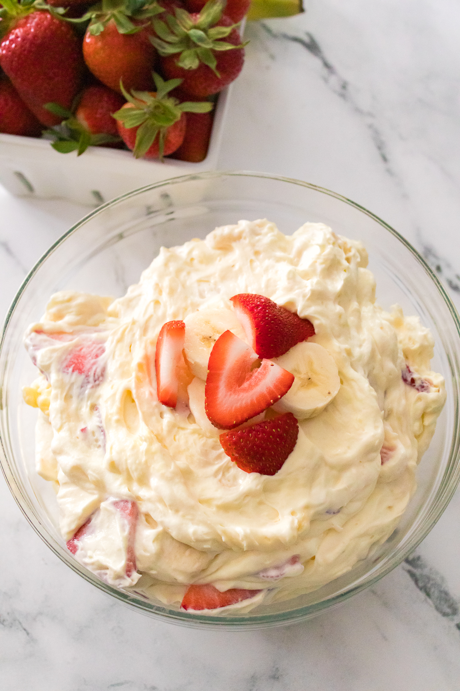 A glass bowl filled with creamy fruit salad topped with sliced strawberries and bananas, with a container of fresh strawberries in the background.