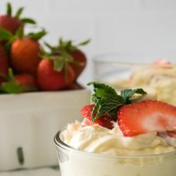 A glass bowl filled with creamy dessert topped with sliced strawberries and a mint leaf, with a container of whole strawberries in the background.