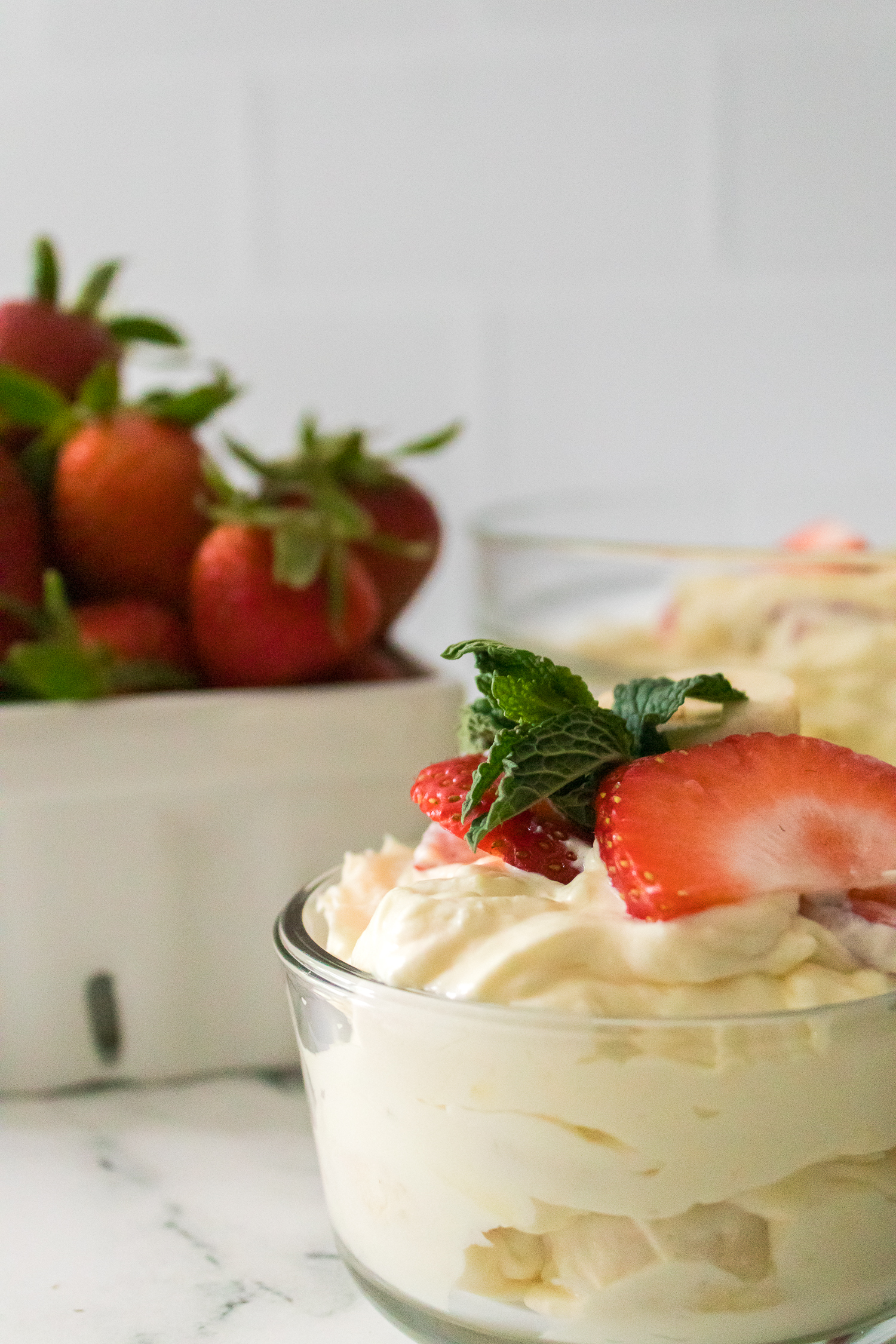 A glass bowl filled with creamy dessert topped with sliced strawberries and a mint leaf, with a container of whole strawberries in the background.