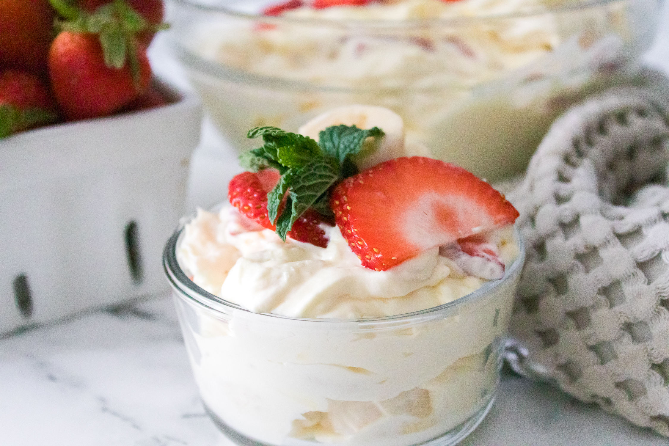 Glass bowl filled with creamy dessert topped with sliced strawberries and mint, with another bowl of the same dessert and a container of strawberries in the background.