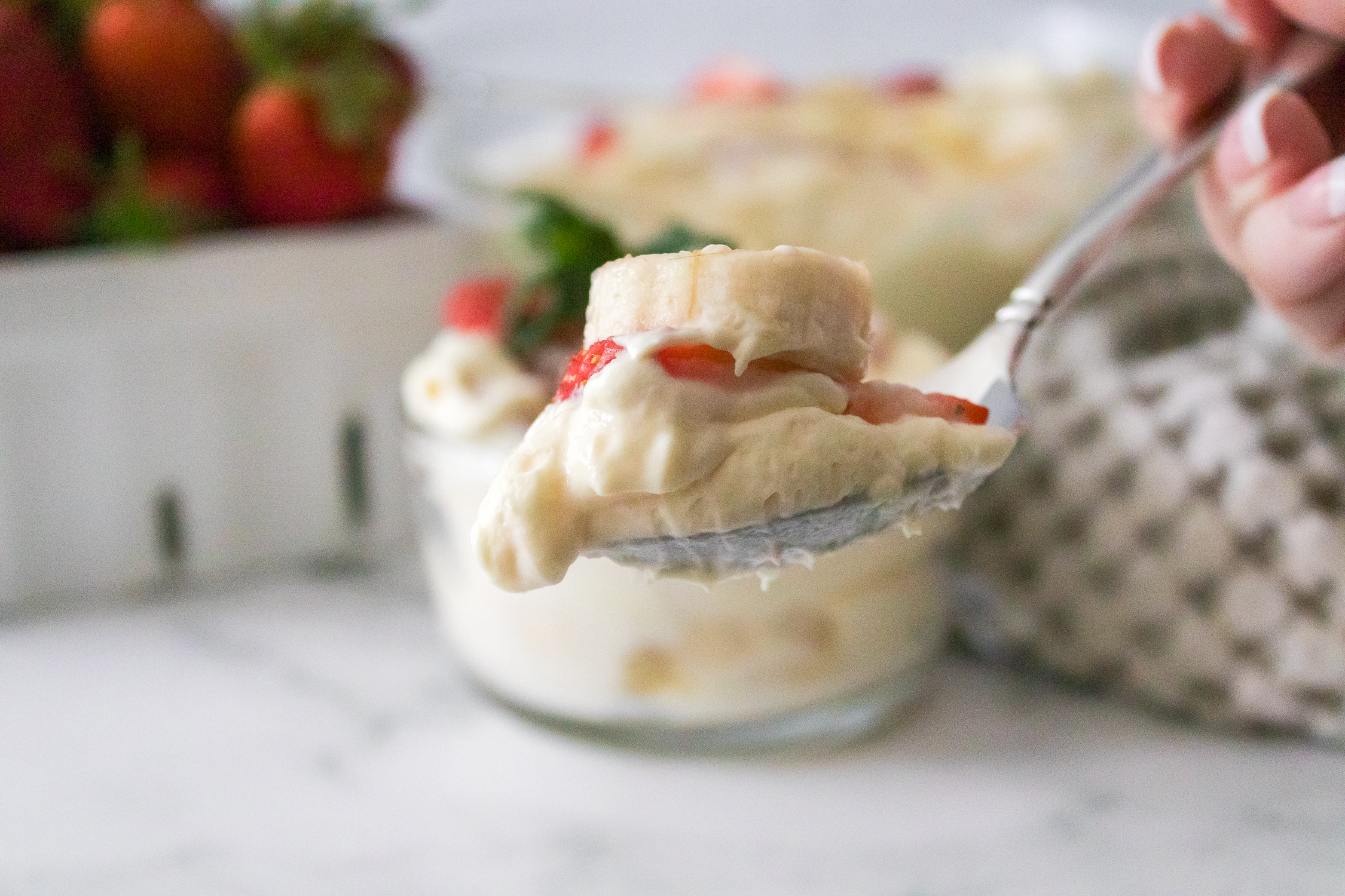 A close-up of a spoonful of Easy strawberry banana pudding dessert with sliced banana and strawberry, held above a bowl of the same dessert on a white surface.  