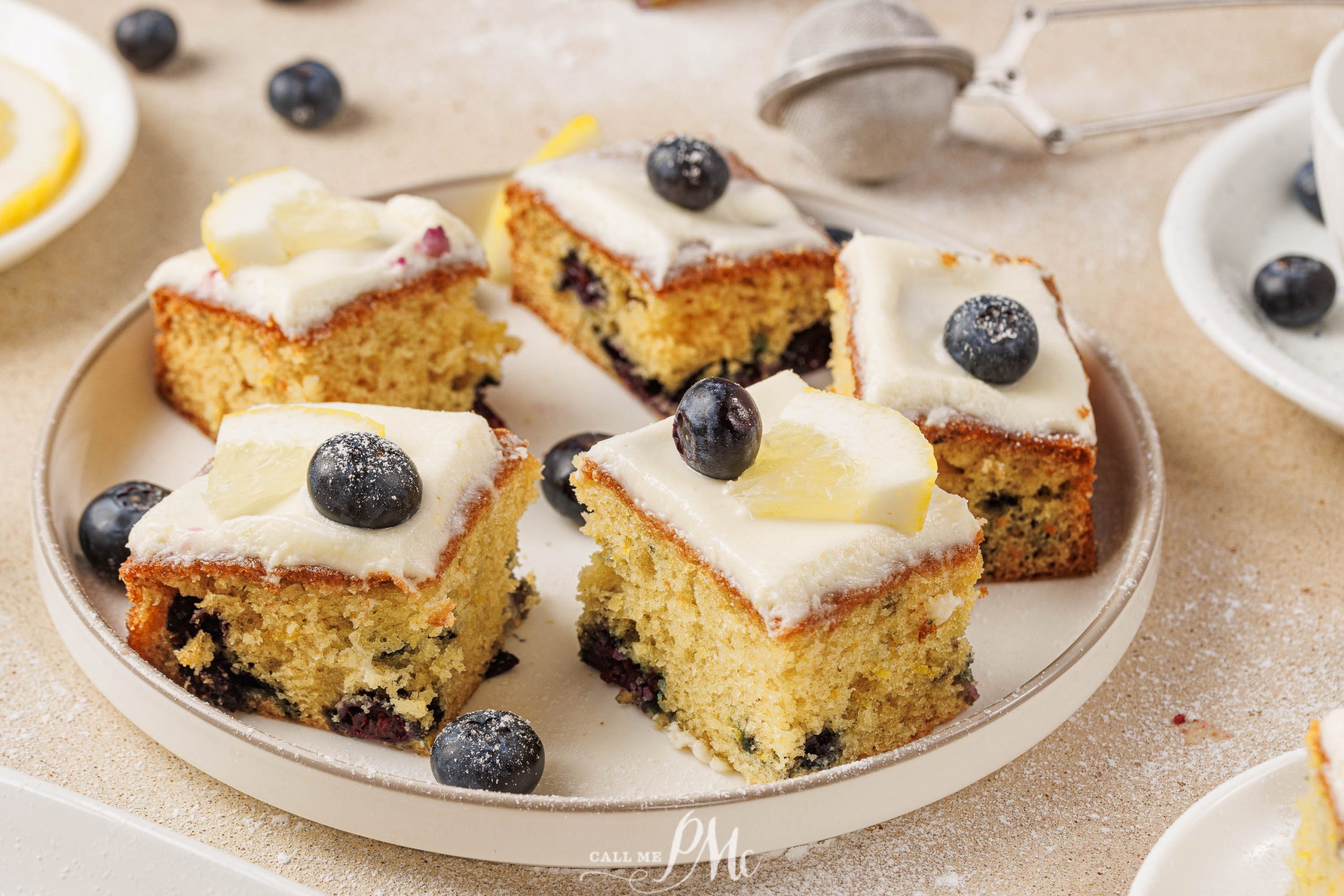 Four pieces of lemon blueberry cake with cream cheese frosting, topped with fresh blueberries and lemon slices, are served on a round plate, with extra blueberries scattered around for a delightful finish.