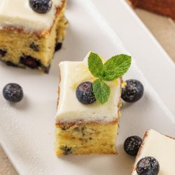 Three pieces of Lemon Blueberry Cake with white frosting are garnished with fresh blueberries and a mint sprig, served on a white rectangular plate.