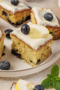 A slice of Lemon Blueberry Cake topped with white icing, a fresh blueberry, and a lemon slice sits on a plate, with more cake pieces in the background.