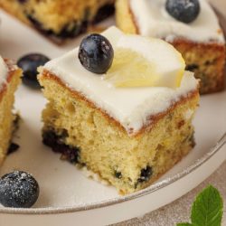 A slice of Lemon Blueberry Cake topped with white icing, a fresh blueberry, and a lemon slice sits on a plate, with more cake pieces in the background.