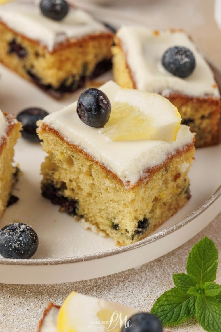 A slice of Lemon Blueberry Cake topped with white icing, a fresh blueberry, and a lemon slice sits on a plate, with more cake pieces in the background.