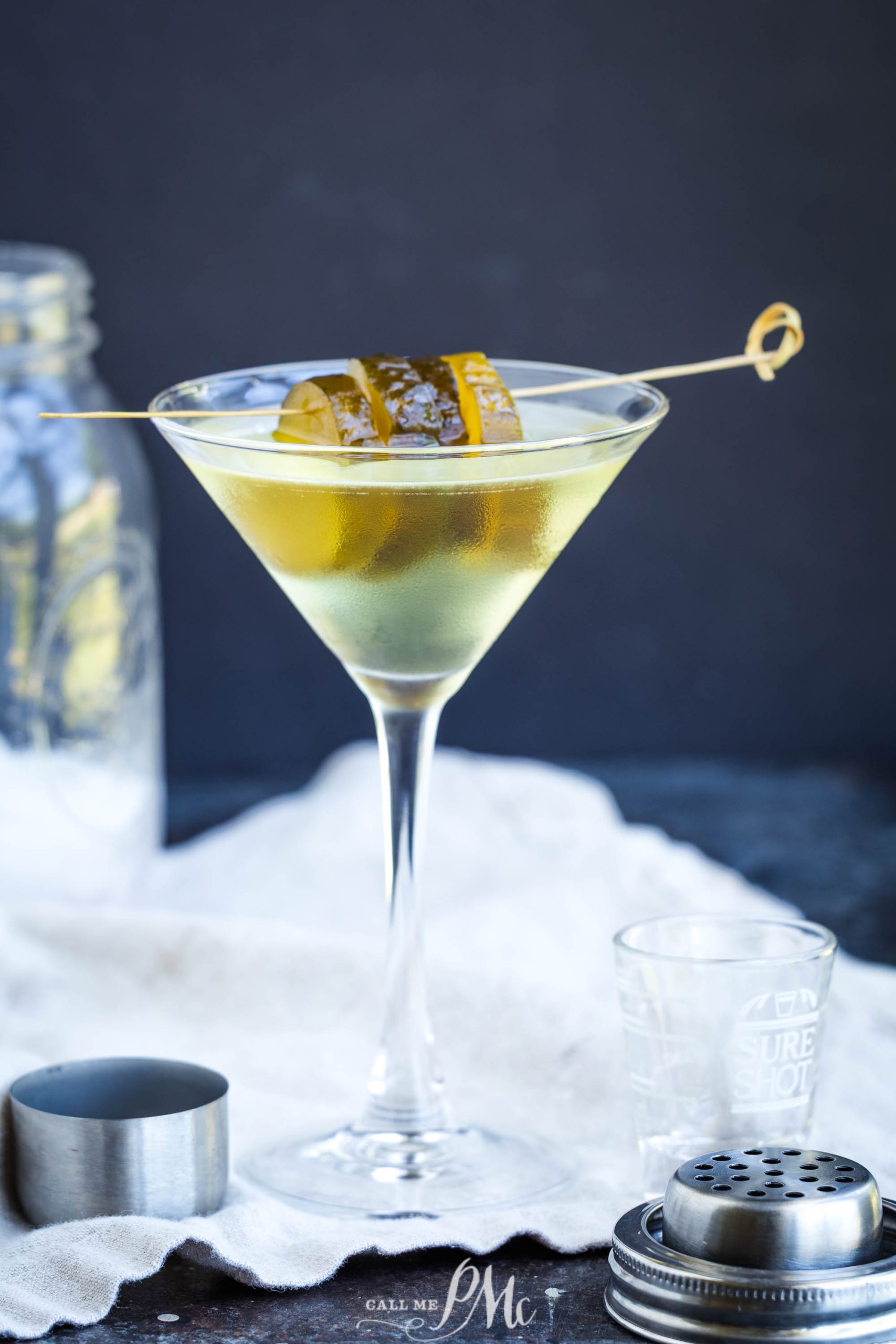 A glass filled with a clear cocktail, garnished with a skewer, sits on a white cloth with a jigger and glass in the background.