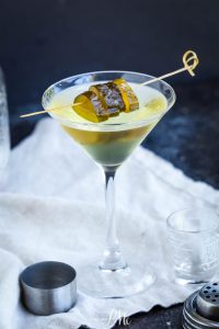 A martini glass filled with a clear yellow-green cocktail, garnished with pickle slices on a wooden skewer, sits on a white cloth with bar tools nearby.